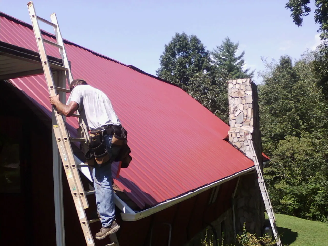 Roofer on a ladder beside a red metal roof and stone chimney — Soffit & Fascia Repair by Roofing Brothers in Delta